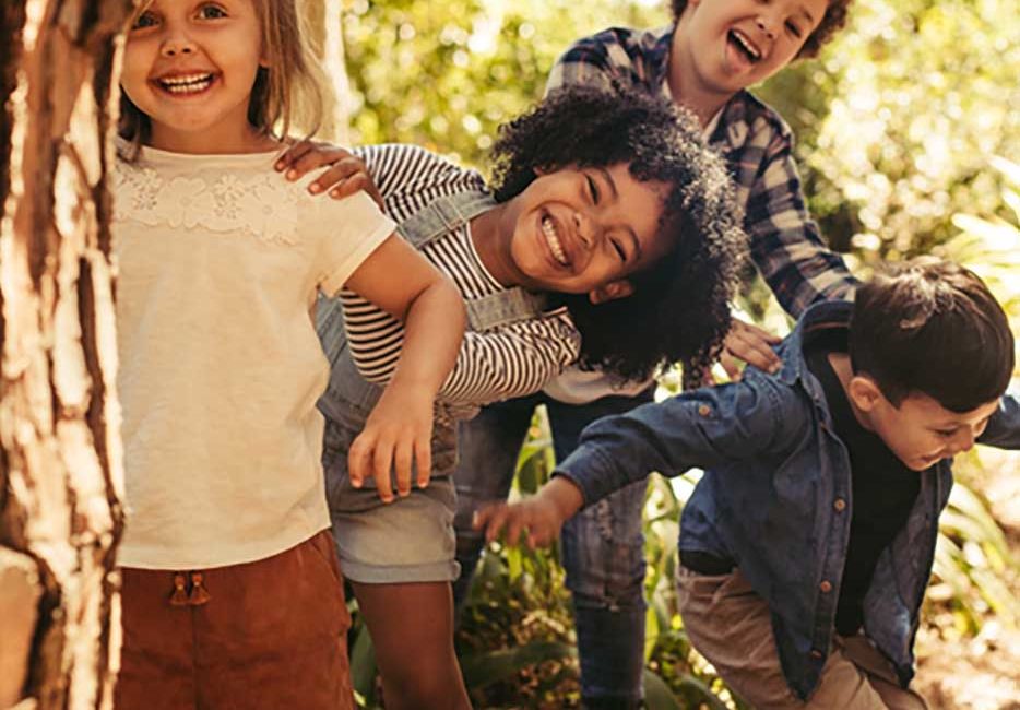 Group of children playing hide and seek Cute smiling kids peeking out from behind the tree in the park. Group of children enjoying playing hide and seek in a forest.