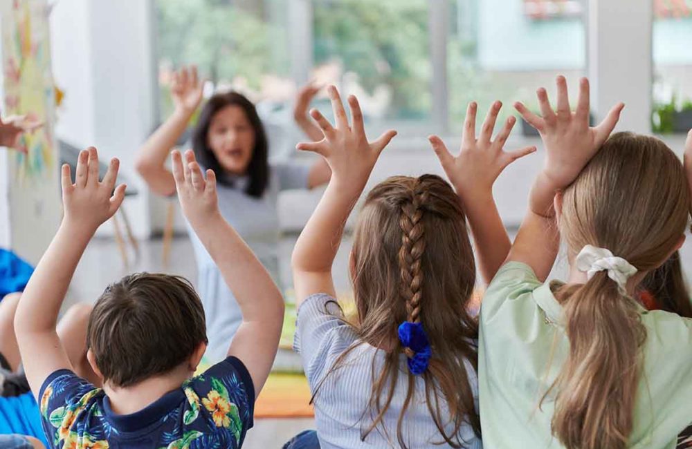 A happy female teacher sitting and playing hand games with a group of little schoolchildren.